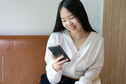 An Asian girl is sitting on brown sofa, reading on her mobile with smile face from Thailand.