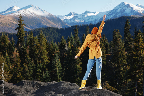Woman full-length hiker in yellow raincoat jumps up with her hands up her back on a mountain trip in the fall and hiking in the mountains in the sunset freedom