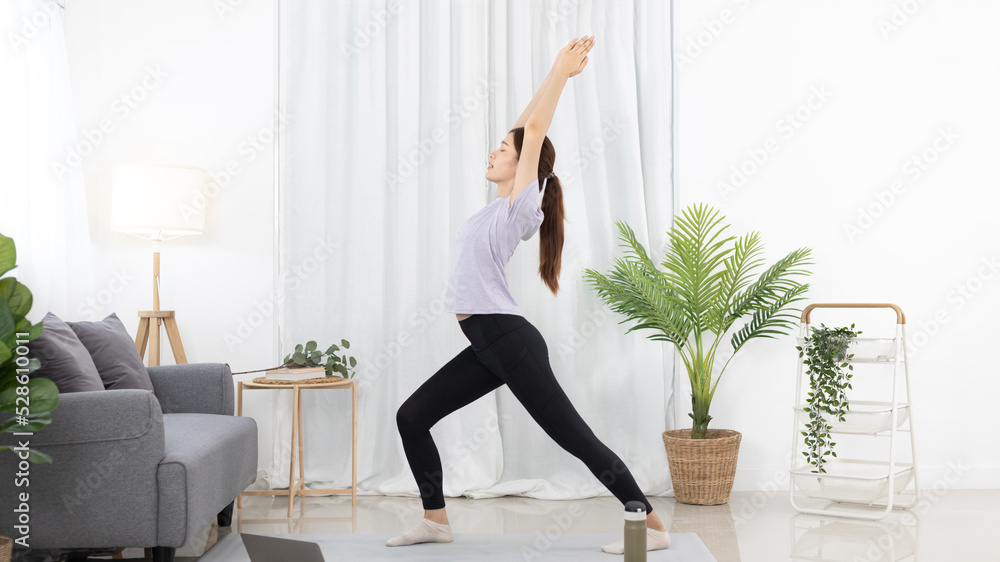 Beautiful Asian woman in sportswear is warming up or doing light exercises in the house with dumbbells and a protein shake or bottle of water and using a laptop at home, Healthy exercise.