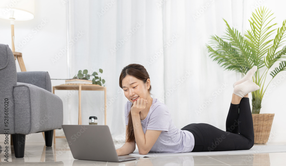 Beautiful Asian woman watching yoga teacher and light exercises in her living room at home, Watch live teaching or VDO on laptop, Dumbbells and a protein shake or bottle of water, Healthy exercise. Beautiful Asian woman watching yoga teacher and light exercises in her living room at home, Watch live teaching or VDO on laptop, Dumbbells and a protein shake or bottle of water, Healthy exercise.