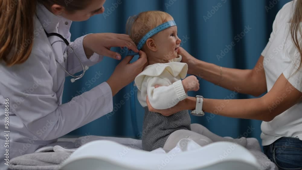 Side view portrait of female toddler sitting in hospital looking up at woman as professional doctor measuring head circumference in slow motion. Carefree Caucasian baby girl on pediatrician visit Side view portrait of female toddler sitting in hospital looking up at woman as professional doctor measuring head circumference in slow motion. Carefree Caucasian baby girl on pediatrician visit