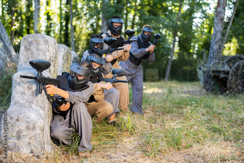 Team taking cover and eliminating opponents on paintball field. People in protective outwear and helmets aiming with paintball markers.