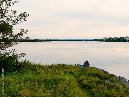 Silhouette of a couple sitting by a lake. Town buildings in the background. Romantic scene with a man and a woman. Love and romance concept. Calm sunset sky.