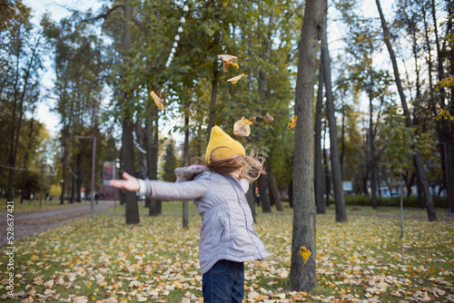 child in demi-season clothes jacket and hat throws up autumn leaves against background of park and trees. kids fun