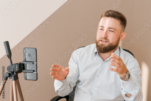 Smiling bearded business man, filming his video blog episode using smartphone, sitting at the office desk writes in a notebook and gesticulates with his hand. Dressed in a shirt photo