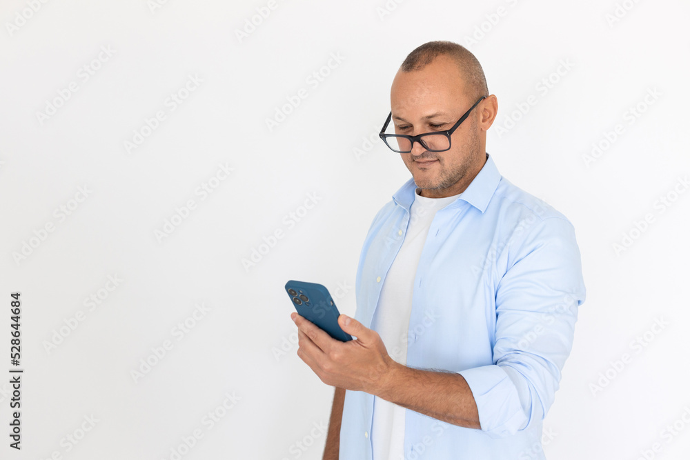 Portrait of happy mature man wearing glasses surfing internet on mobile phone. Balding Caucasian businessman wearing white T-shirt and blue shirt using smartphone. Mobile communication concept Portrait of happy mature man wearing glasses surfing internet on mobile phone. Balding Caucasian businessman wearing white T-shirt and blue shirt using smartphone. Mobile communication concept