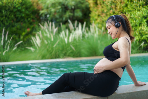 pregnant woman sitting on edge of swimming pool while listening to music with headphone © geargodz