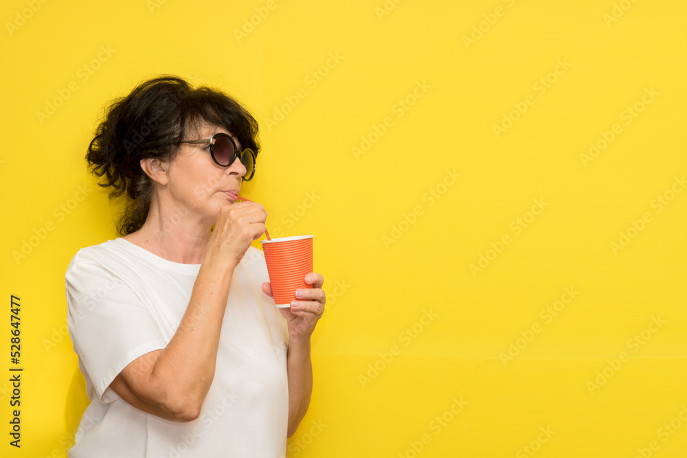stylish aged woman tilting her head drinks from a paper glass with a straw on a yellow background with copy space stylish aged woman tilting her head drinks from a paper glass with a straw on a yellow background with copy space
