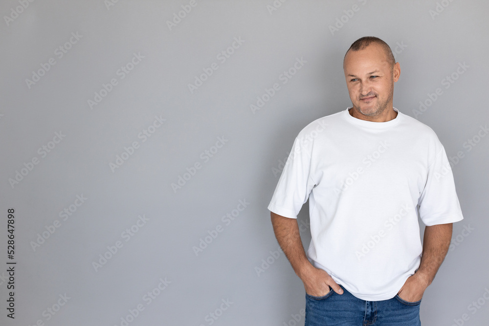 Portrait of smiling mature man wearing white T-shirt and jeans. Balding Caucasian man posing against gray background. Masculinity concept Portrait of smiling mature man wearing white T-shirt and jeans. Balding Caucasian man posing against gray background. Masculinity concept