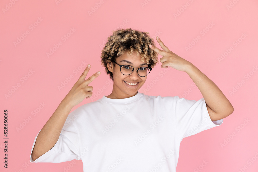 Portrait of happy mixed race woman making peace gesture. Young woman wearing eyeglasses and white T-shirt looking at camera and smiling against pink background. Success concept Portrait of happy mixed race woman making peace gesture. Young woman wearing eyeglasses and white T-shirt looking at camera and smiling against pink background. Success concept