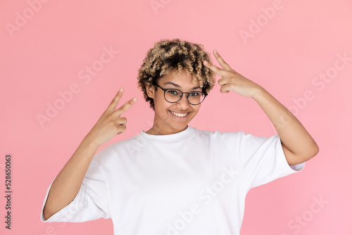 Portrait of happy mixed race woman making peace gesture. Young woman wearing eyeglasses and white T-shirt looking at camera and smiling against pink background. Success concept