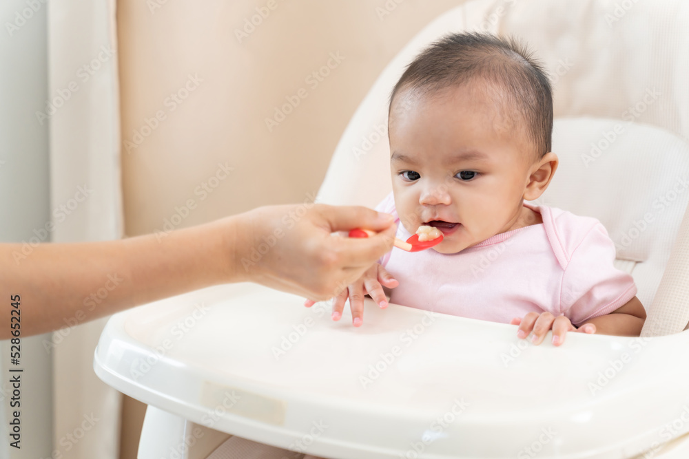 Asian attractive parents feeding foods to baby toddler in bedroom. Happy family,loving mother cook and serve healthy foods to small child sits on a chair and eating with spoon for lunch at home Asian attractive parents feeding foods to baby toddler in bedroom. Happy family,loving mother cook and serve healthy foods to small child sits on a chair and eating with spoon for lunch at home