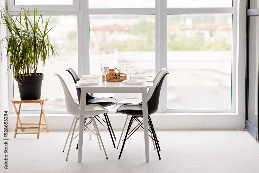 Fototapeta premium Modern interior of a cozy dining room kitchen, white furniture, against the background of a panoramic window. Conceptual decor design, house mortgage loan for a young family. Soft selective focus.