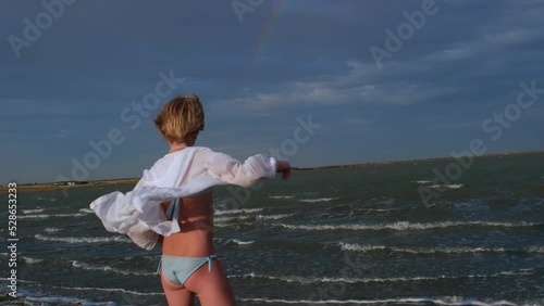 A beautiful teenage girl dances with pleasure on the shore of the sea or lake. A child in a swimsuit stands with his back to the camera in the sun. A girl in a white unbuttoned shirt moves towards the