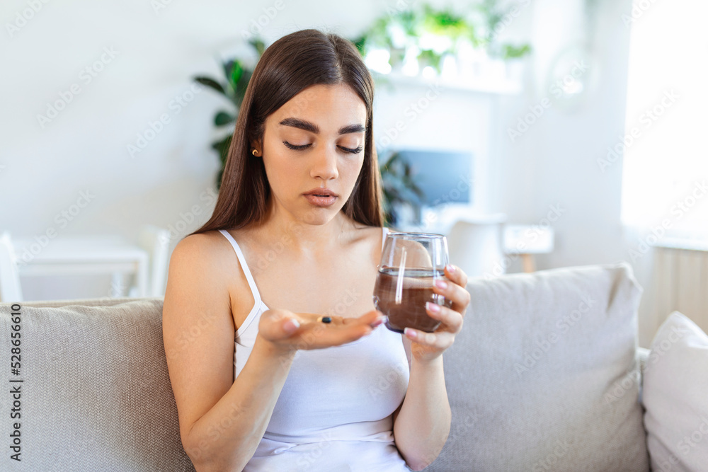 Concept of suffering from terrible pain. Beautiful, sad, upset, unhappy, troubled, weak woman with black hair, wearing casual clothes is sitting on a sofa and taking a pill Concept of suffering from terrible pain. Beautiful, sad, upset, unhappy, troubled, weak woman with black hair, wearing casual clothes is sitting on a sofa and taking a pill