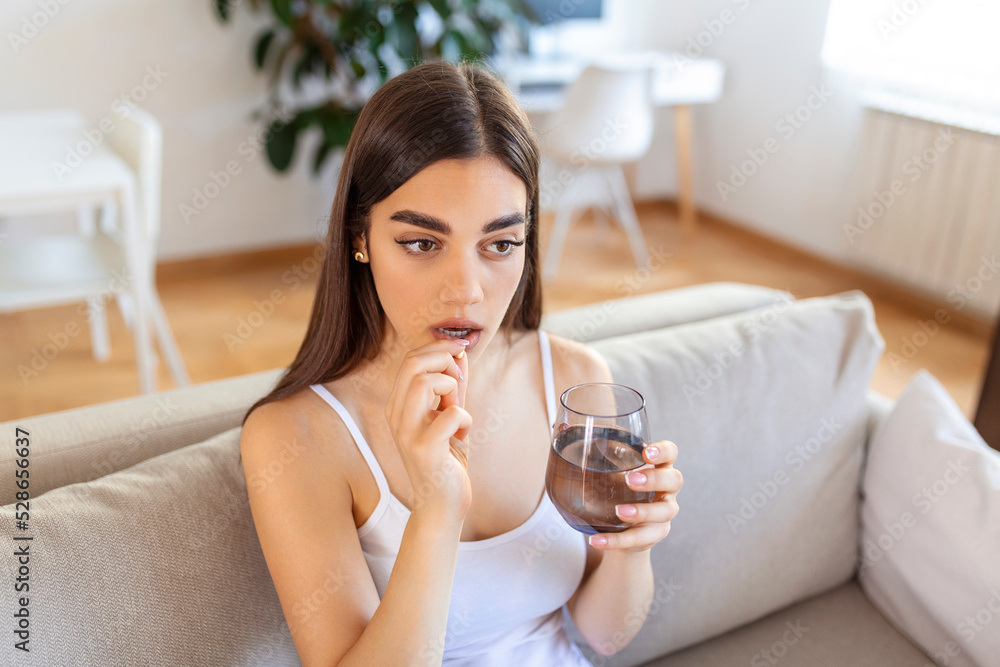 Concept of suffering from terrible pain. Beautiful, sad, upset, unhappy, troubled, weak woman with black hair, wearing casual clothes is sitting on a sofa and taking a pill Concept of suffering from terrible pain. Beautiful, sad, upset, unhappy, troubled, weak woman with black hair, wearing casual clothes is sitting on a sofa and taking a pill