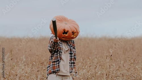 A funny woman in a shirt with the head of a large pumpkin holds a bat in her hands, grimaces at the camera and threatens outdoors in a field. Halloween concept