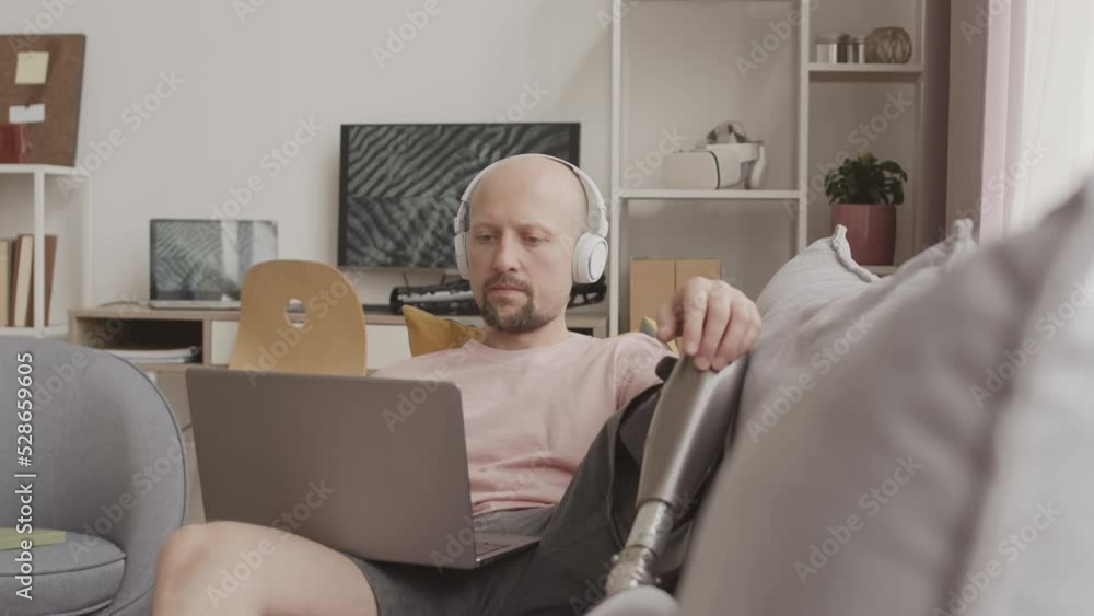 Slowmo of young Caucasian man with prosthetic leg listening to music in headphones while texting with friends on laptop, resting on couch in his apartment Slowmo of young Caucasian man with prosthetic leg listening to music in headphones while texting with friends on laptop, resting on couch in his apartment