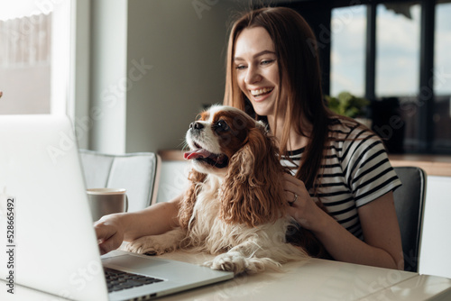 Delightful smiling young woman sitting in kitchen with dog coker spaniel, use laptop. Video call to friends and family