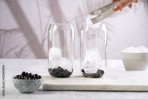 Female hand putting ice into a transparent glass with cooked tapioca pearls for trendy bubble boba ice tea in two small grey ceramic bowls on marble board on dark grey concrete background