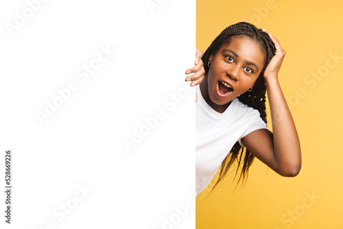 Valokuva Surprised dark skinned woman looking at empty white banner, hiding behind huge p