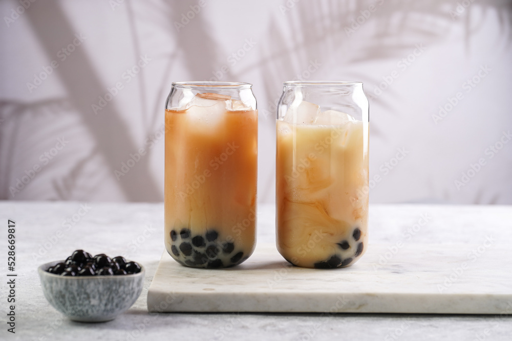 Two glasses with black tea, milk, ice cubes and cooked tapioca pearls for trendy bubble boba ice tea, two small grey ceramic bowls on marble board on pastel tropical background Two glasses with black tea, milk, ice cubes and cooked tapioca pearls for trendy bubble boba ice tea, two small grey ceramic bowls on marble board on pastel tropical background