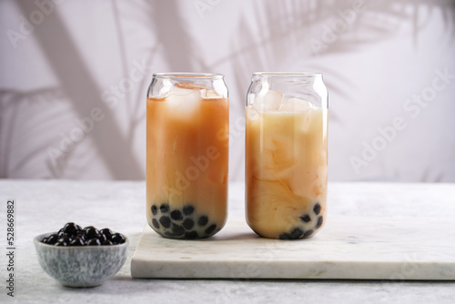 Two glasses with black tea, milk, ice cubes and cooked tapioca pearls for trendy bubble boba ice tea, two small grey ceramic bowls on marble board on pastel tropical background