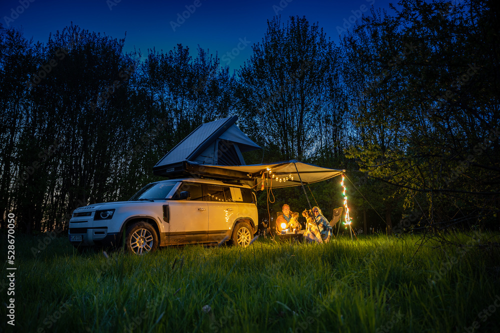 Man and women with dogs camping in front of a 4x4 Offroad vehicle with roof tent at night time and romantic lighting Man and women with dogs camping in front of a 4x4 Offroad vehicle with roof tent at night time and romantic lighting