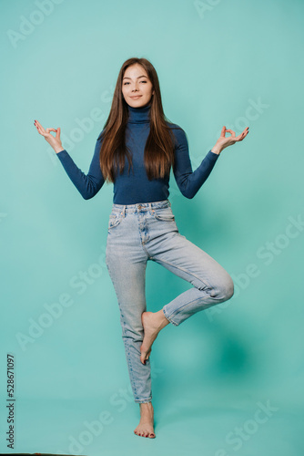Vertical shoot of beautiful calm young woman with loose hair standing in yoga pose over turquoise background. Attractive Caucasian girl posing at studio dressed in casual. Balance concept.