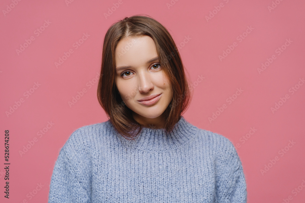 Close up shot of satisfied dark haired young lady tilts head, enjoys lovely casual talk, wears warm winter sweater, poses alone against pink background, gazes focused at camera, feels relaxed Close up shot of satisfied dark haired young lady tilts head, enjoys lovely casual talk, wears warm winter sweater, poses alone against pink background, gazes focused at camera, feels relaxed