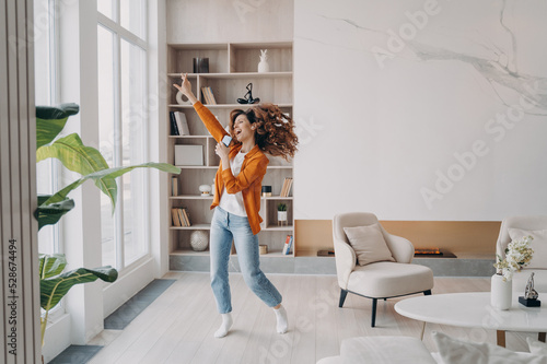 Energetic woman holding smartphone, having fun, dances to music rejoicing with good news at home