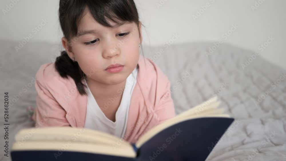 Close up of little girl in pink sweatshirt laying on the bed reading book. Child is turning the pages with an interest. Study at home, Homeschooling concept and cozy leisure time. Close up of little girl in pink sweatshirt laying on the bed reading book. Child is turning the pages with an interest. Study at home, Homeschooling concept and cozy leisure time.