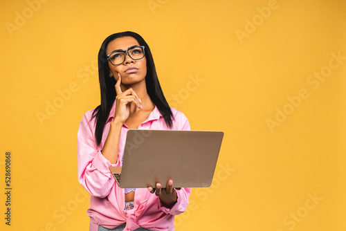 Young african american black thinking serious lady using laptop and smiling isolated over yellow background.