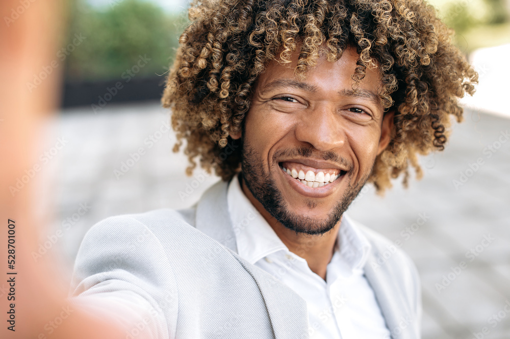 Cheerful handsome curly haired mixed race man standing outdoors, taking a selfie on his smartphone, mixed race male blogger having live streaming on social network, smiling. Selfie photo concept Cheerful handsome curly haired mixed race man standing outdoors, taking a selfie on his smartphone, mixed race male blogger having live streaming on social network, smiling. Selfie photo concept
