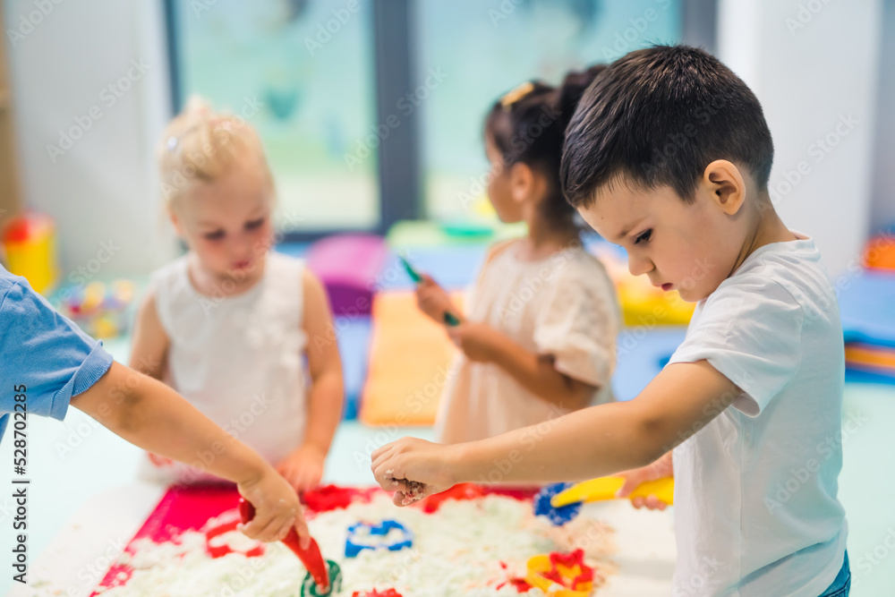 Relaxing sensory play with moldable kinetic sand at nursery school. Toddlers with their teacher having fun around the table using different tools for sculpting sand such as colorful and textured Relaxing sensory play with moldable kinetic sand at nursery school. Toddlers with their teacher having fun around the table using different tools for sculpting sand such as colorful and textured