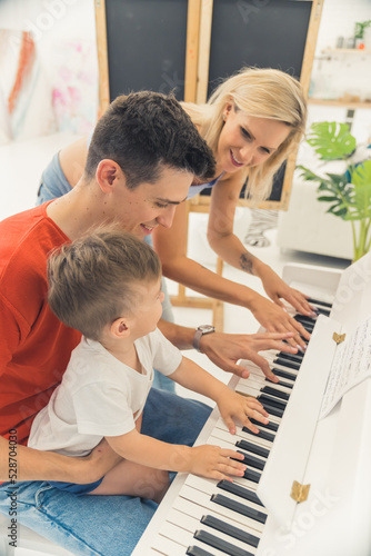 Vertical indoor shot of a happy family of three - heterosexual caucasian married couple and their cute preschooler son - sitting by the piano and playing. Fun music entertainment for children. High
