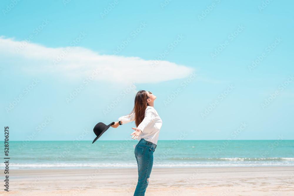 A beautiful girl wearing a white dress with a black hat stands alone and open arms on the beach by the sea on a clear day. She was glad to come to the sea on vacation.