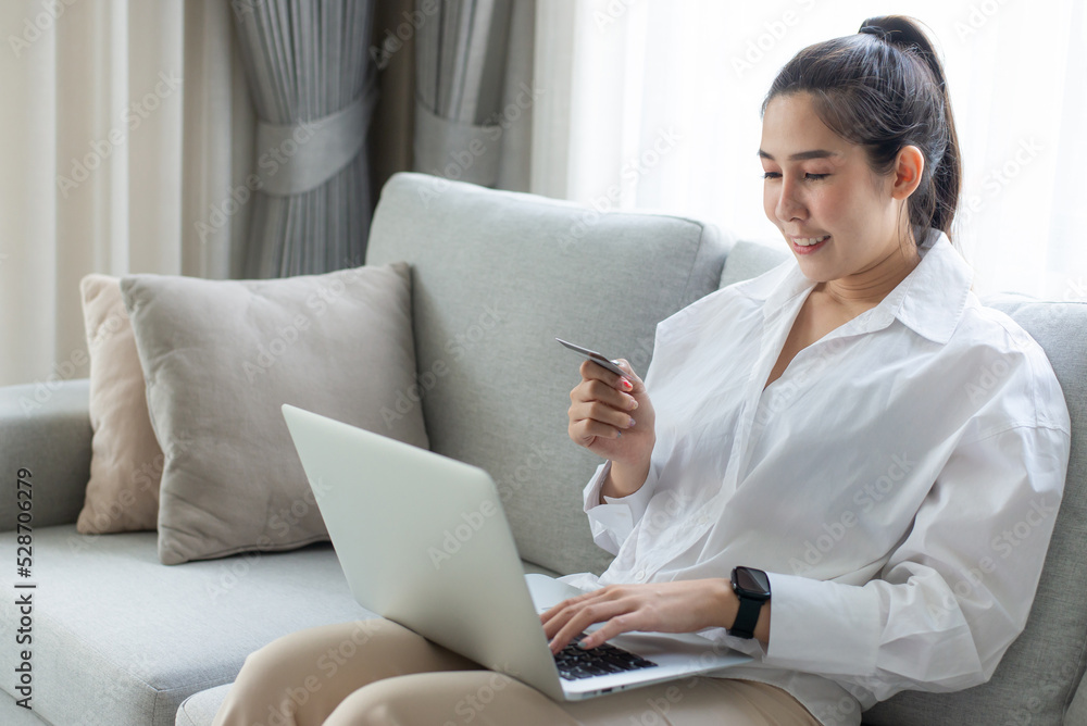 A woman in a white shirt sits and using a computer laptop and credit card for pay online shopping on the couch at home. She smiled happily not having to leave the house to go shopping. A woman in a white shirt sits and using a computer laptop and credit card for pay online shopping on the couch at home. She smiled happily not having to leave the house to go shopping.
