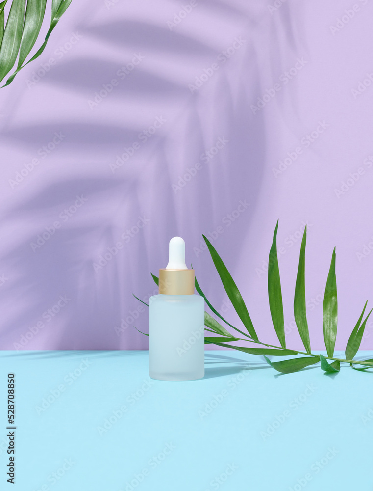 A white glass bottle with a pipette and a green palm leaf. Purple background. A white glass bottle with a pipette and a green palm leaf. Purple background.