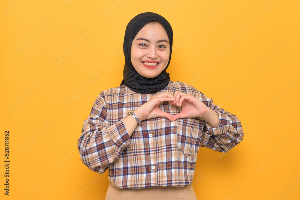 Smiling young Asian woman in plaid shirt showing heart gesture and looking at camera isolated on yellow background Smiling young Asian woman in plaid shirt showing heart gesture and looking at camera isolated on yellow background