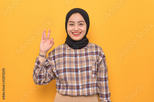 Cheerful young Asian woman in plaid shirt showing okay gesture demonstrates symbol of approval isolated on yellow background