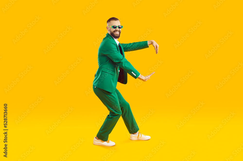Funny young guy dancing in a cool party outfit. Happy, joyful man wearing a stylish green suit and sunglasses dancing isolated on a bright yellow colour background. Full body studio shot, side view Funny young guy dancing in a cool party outfit. Happy, joyful man wearing a stylish green suit and sunglasses dancing isolated on a bright yellow colour background. Full body studio shot, side view