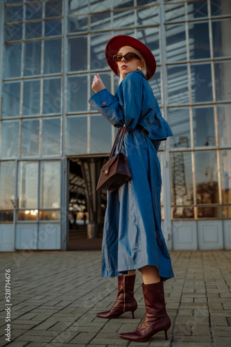 Fashionable confident blonde woman wearing dark red hat, trendy sunglasses, blue midi trench coat, leather ankle boots, holding stylish bag, posing in street of city. Outdoor full-length portrait