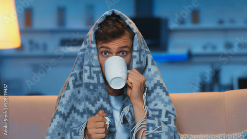 sick man with blue eyes drinking tea while sitting under blanket in living room photo