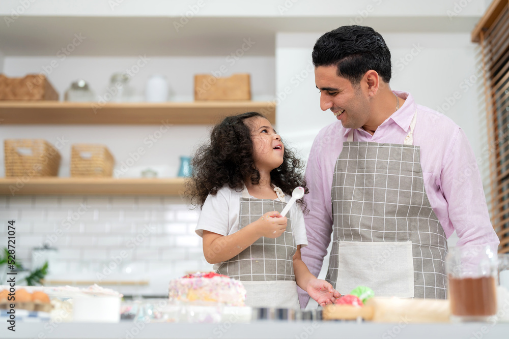 Young little girl standing at counter baking bakery in the modern kitchen at home. Happy smiling and enjoy on weekend. Kids and father wearing apron and smile together. Homemade bakery concept Young little girl standing at counter baking bakery in the modern kitchen at home. Happy smiling and enjoy on weekend. Kids and father wearing apron and smile together. Homemade bakery concept