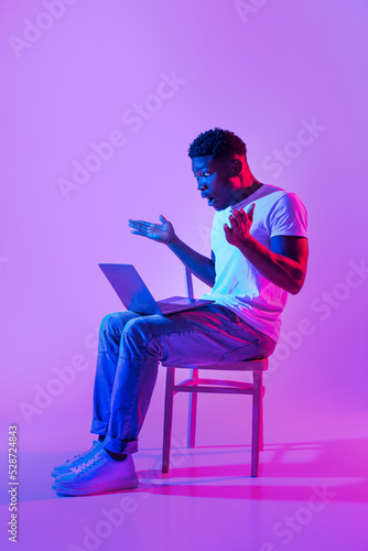 Emotional young black man sitting on chair with laptop, feeling shocked, making mistake in business project in neon light