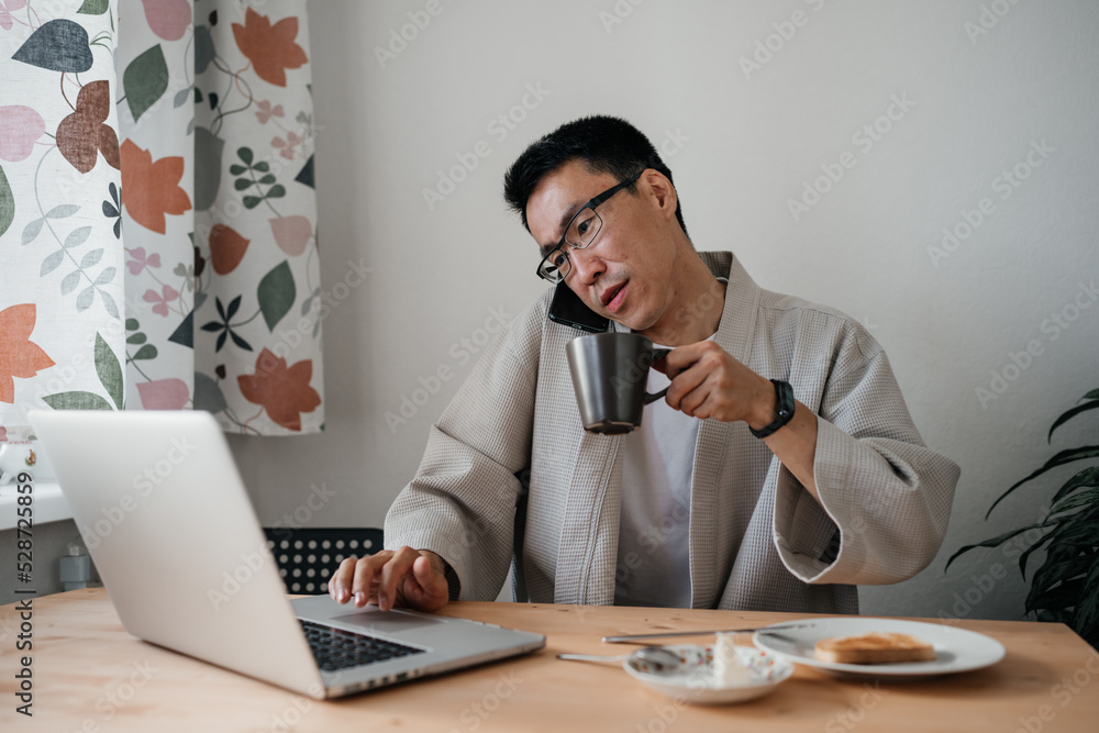 Middle aged asian man in spectacles with smartphone and laptop in kitchen.