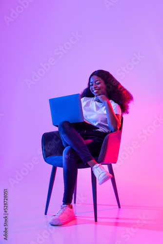 Joyful young black lady sitting in armchair with modern laptop, working remotely or video chatting in neon light, full length