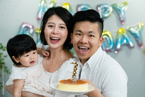 joyful asian father and mother celebrating first birthday of their baby daughter with a cake while smiling at the camera at home with festive decoration