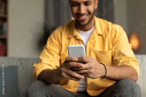 Happy young black male using smartphone, browsing internet, posting in social media at home, selective focus